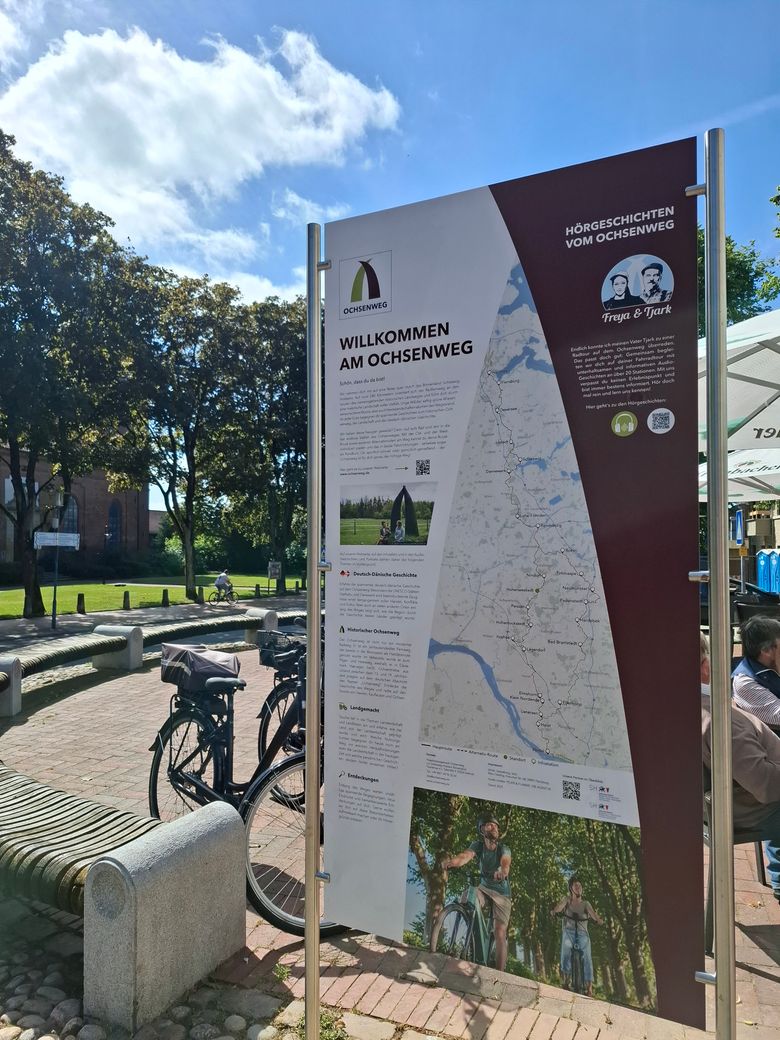 Eine Infotafel in Hohenwestedt am Ochsenweg. Im Hintergrund sind Bäume und Fahrräder zu sehen. Der Himmel ist blau mit einigen Wolken.
