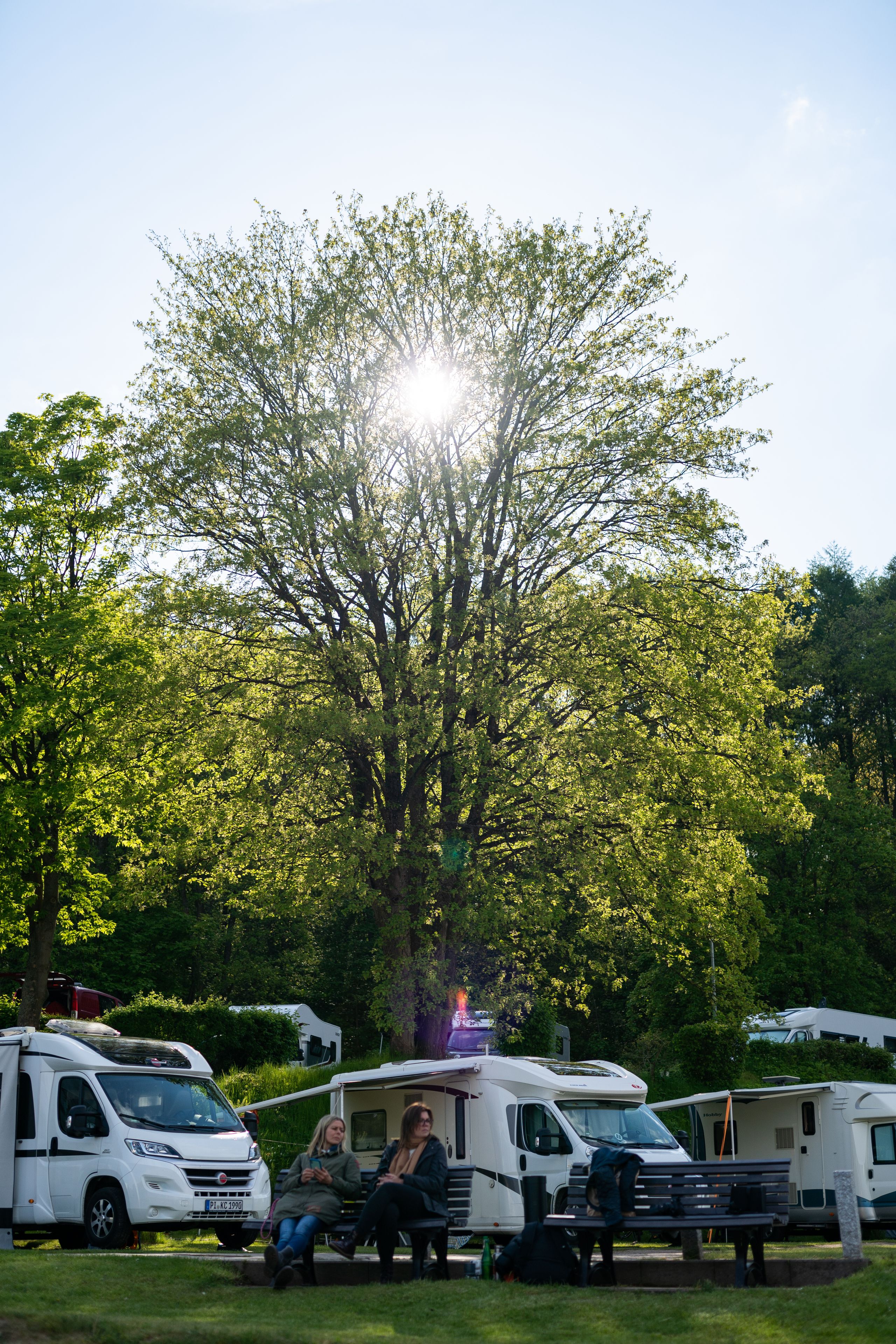 Zwei Personen entspannen auf einer Bank vor Wohnmobilen, umgeben von grünen Bäumen und strahlendem Sonnenschein.