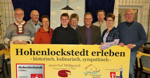 Acht Personen präsentieren das Banner „Hohenlockstedt erleben“ in einem Innenraum.