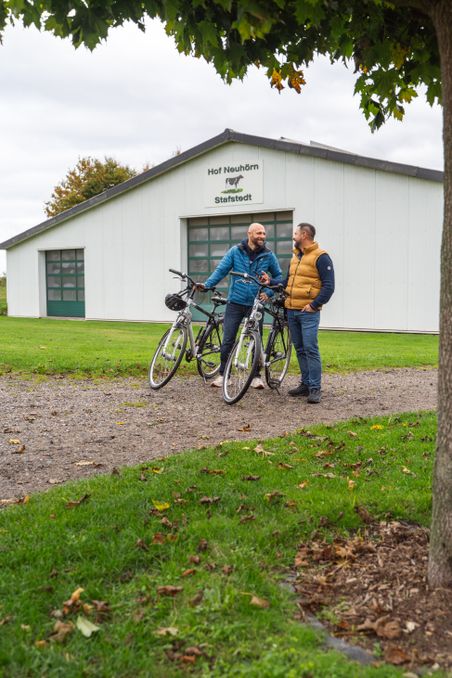 Zwei Männer stehen mit Fahrrädern vor dem Gebäude 'Hof Neuhörn Stafstedt'. Sie unterhalten sich unter einem Baum.