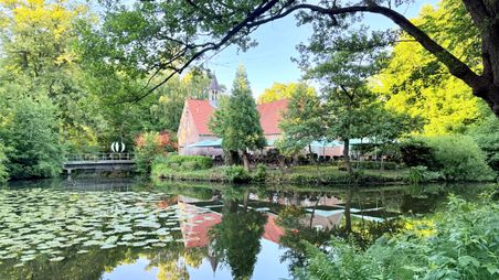 Ein idyllisches Gebäude mit rotem Dach spiegelt sich im Mühlenteich, umgeben von üppigem Grün und Seerosen.