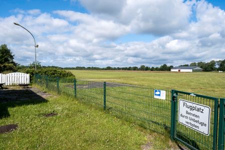 Ein umzäunter Flugplatz mit einem Verbotsschild für Unbefugte. Im Hintergrund erstreckt sich eine grüne Wiese und ein Gebäude.