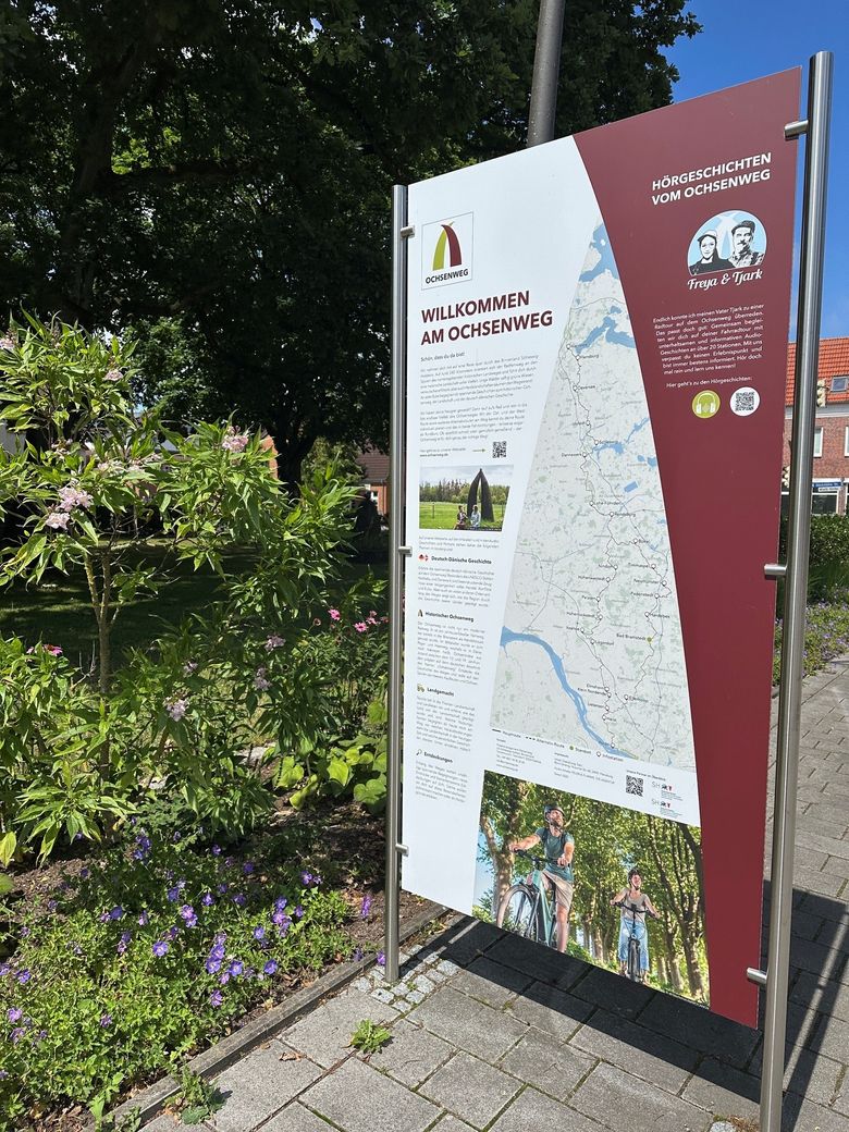 Eine Infotafel in Bad Bramstedt am Ochsenweg zeigt eine Karte und Text über die Route. Es steht auf einem gepflasterten Weg, umgeben von grünen Pflanzen und Blumen.