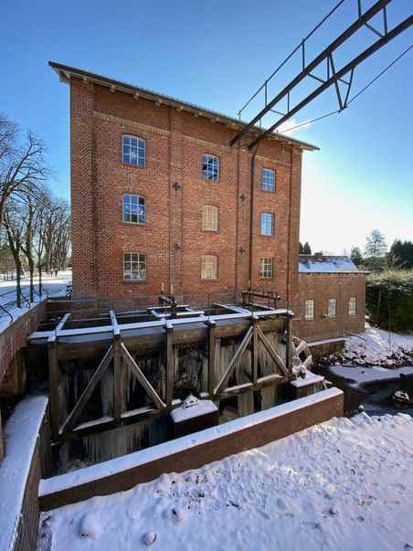 Ein Backsteingebäude mit einem gefrorenen Mühlenrad im Vordergrund, umgeben von Schnee und klarem Himmel.