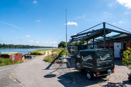 Brauers Aalkate liegt idyllisch am Flussufer. Ein kleiner Lieferwagen steht vor dem Restaurant, das von grüner Natur umgeben ist.