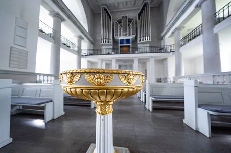 Ein kunstvoll verziertes Taufbecken in der Vicelinkirche, umgeben von weißen Kirchenbänken und einer großen Orgel im Hintergrund.
