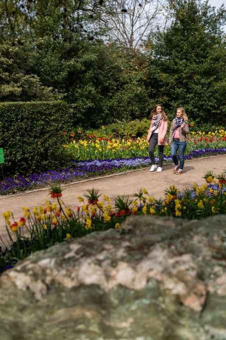 Zwei Frauen spazieren im Holstein Arboretum Ellerhoop zwischen bunten Blumenbeeten und hohen Bäumen.