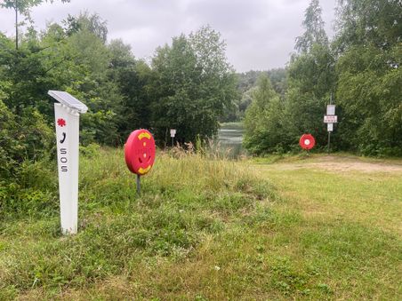 Grüner Uferbereich mit SOS-Notrufpfosten und Rettungsringen. Im Hintergrund ist ein See von Bäumen umgeben.