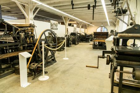 Historische Druckmaschinen im Druckmuseum Rendsburg, aufgereiht in einem großen Raum mit Holzbalkendecke.