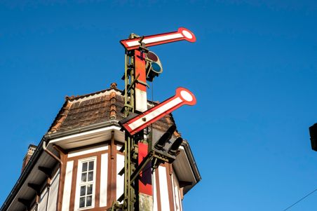 Ein historisches Stellwerk mit roten Signalen vor einem klaren blauen Himmel. Das Gebäude hat ein markantes Dach und weiße Fenster.