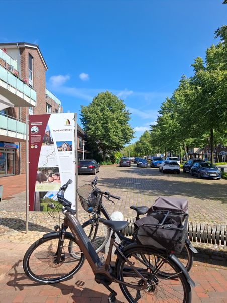 Zwei Fahrräder stehen vor einer Infotafel in Hohenwestedt. Im Hintergrund sind Bäume, Autos und ein Gebäude zu sehen.