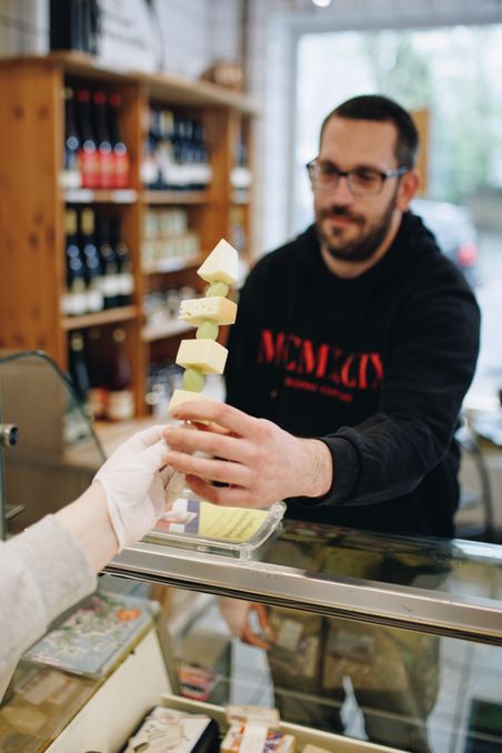 Ein Verkäufer überreicht einem Kunden einen Spieß mit Käse und Trauben in einem Feinkostladen.
