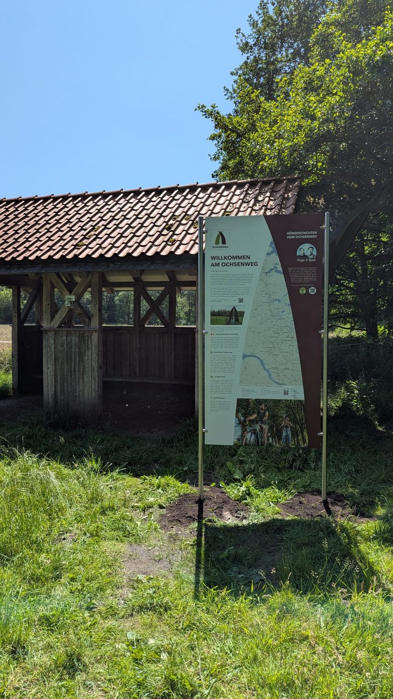 Ein Schild am Ochsenweg informiert Besucher vor einem hölzernen Unterstand mit Ziegeldach. Der Himmel ist klar und sonnig.