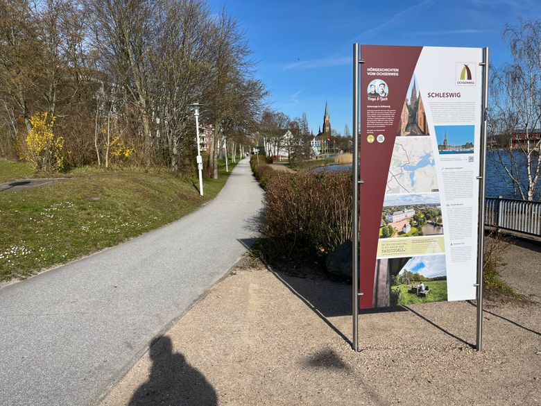 Ein Informationsschild am Ochsenweg in Schleswig. Der Weg führt an einem See entlang, umgeben von Bäumen und Sträuchern unter blauem Himmel.