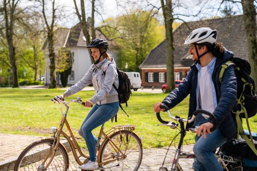 Zwei Personen fahren lächelnd mit Fahrrädern durch einen sonnigen Park. Sie tragen Helme und Rucksäcke, während sie sich unterhalten.