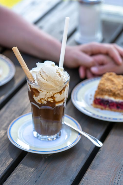 Eiskaffee mit Vanilleeis und Sahne auf einem Unterteller, daneben Dessert und Hände am Tisch.