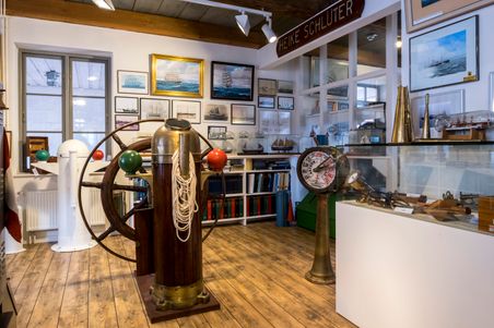 Ein Raum im Schifffahrtsarchiv Rendsburg zeigt nautische Instrumente, Schiffsmodelle und historische Bilder. Ein Steuerrad und ein Maschinentelegraf sind zu sehen.