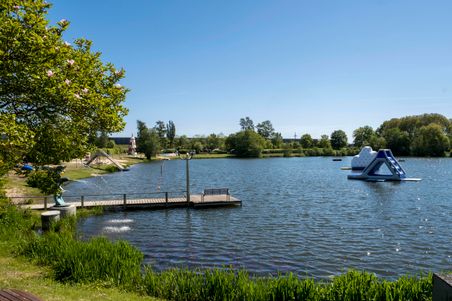 Ein idyllischer See mit Steg und Wasserspielgeräten. Im Hintergrund ein Spielplatz und ein Leuchtturm. Perfekt für Familienausflüge.