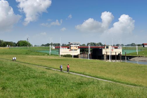 Radfahrer fahren auf einem Weg entlang einer grünen Wiese neben dem Krückau-Sperrwerk. Der Himmel ist blau mit einigen Wolken.