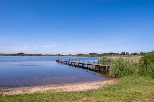 Ein Holzsteg ragt in den ruhigen Vollstedter See, umgeben von Schilf und grünen Wiesen. Der Himmel ist klar und blau.