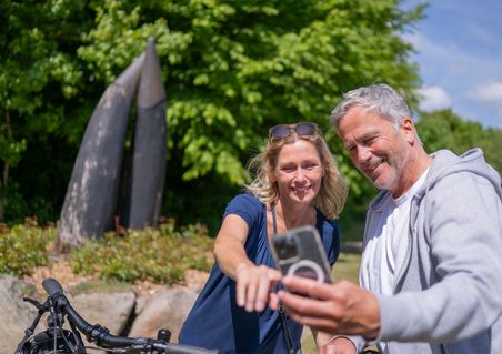Ein Mann und eine Frau machen ein Selfie vor einer Skulptur im Freien. Im Hintergrund sind Bäume und ein sonniger Himmel zu sehen.