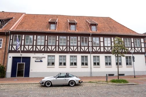 Das Schifffahrtsarchiv Rendsburg in einem historischen Fachwerkgebäude. Ein silbernes Auto ist davor geparkt. Ein Baum und eine Laterne stehen daneben.