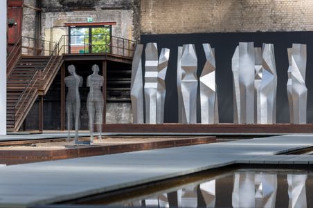 Moderne Skulpturen in einer Industriehalle. Zwei filigrane Figuren stehen vor einer Reihe abstrakter, metallischer Formen. Ein Wasserbecken spiegelt die Szene.