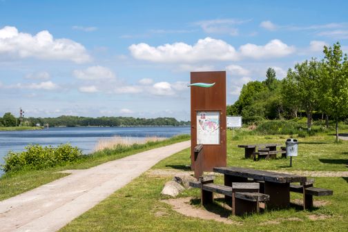 Schiffsanleger in Schacht-Audorf mit Picknicktischen und einem Informationsschild am Flussufer. Der Himmel ist blau mit einigen Wolken.