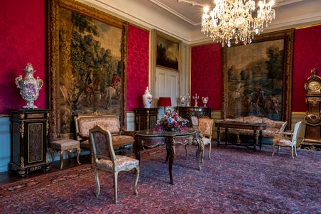 Eleganter Salon im Herrenhaus Emkendorf mit antiken Möbeln, Wandteppichen und einem Kronleuchter. Die Wände sind in Rot gehalten, der Raum wirkt luxuriös.