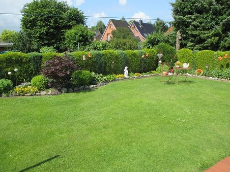 Gepflegter Garten mit großem Rasen, Hecken, Blumenbeeten und Dekofiguren; im Hintergrund stehen Wohnhäuser.