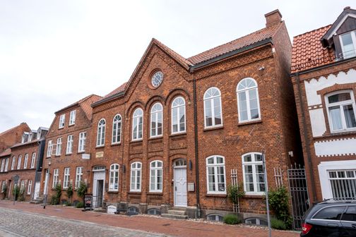 Ein historisches Backsteingebäude mit weißen Fenstern und einem Schild, das auf ein Museum hinweist. Es befindet sich in einer gepflasterten Straße.