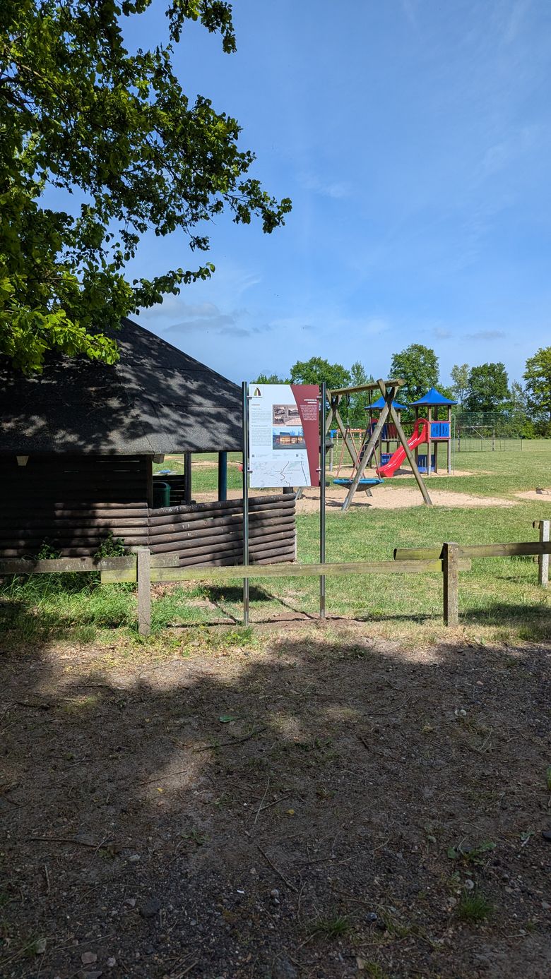 Ein Spielplatz mit Schaukeln und Rutsche neben einer Holzhütte. Eine Informationstafel steht im Vordergrund. Der Himmel ist blau und Bäume umgeben die Szene.