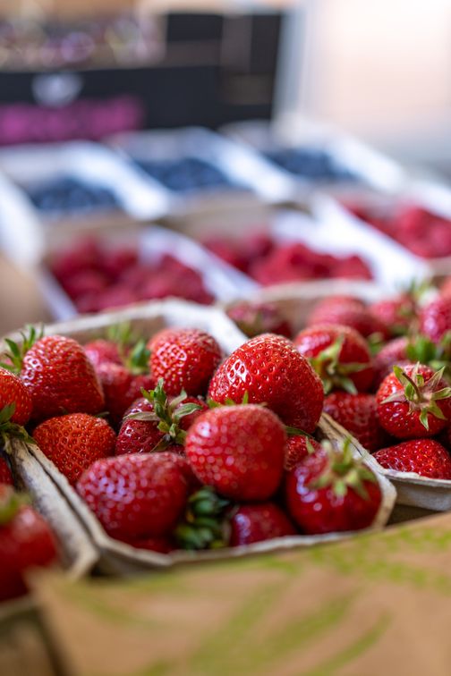 Frische Erdbeeren in einem Karton, im Vordergrund eines Marktstands. Im Hintergrund sind weitere Beeren in Schalen zu sehen.