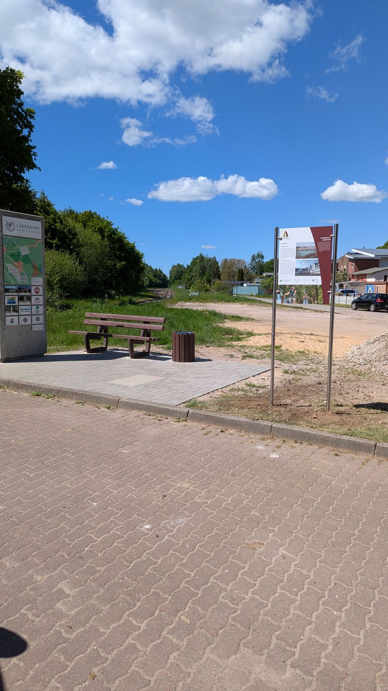 Ein Rastplatz in Lägerdorf mit einer Bank und Infotafeln. Im Hintergrund sind Bahngleise und ein Auto zu sehen. Der Himmel ist blau mit Wolken.