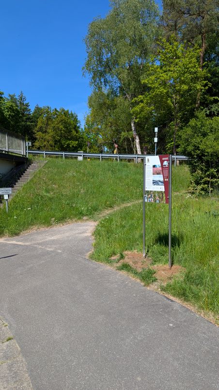 Ein Weg mit einem Informationsschild und einer Treppe, umgeben von grüner Vegetation und einem blauen Himmel.