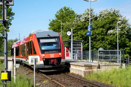 Ein Regionalzug der Deutschen Bahn hält am Bahnhof Schülldorf. Der Bahnsteig ist von grünen Bäumen umgeben und eine Uhr zeigt die Uhrzeit an.