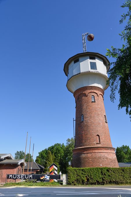 Roter Backsteinturm mit weißem Aufbau und Kugel auf dem Dach vor blauem Himmel; unten ein Museumsschild.