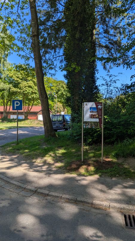 Eine Informationstafel steht am Wegesrand. Im Hintergrund sind Bäume und ein geparktes Auto zu sehen. Der Himmel ist klar und blau.