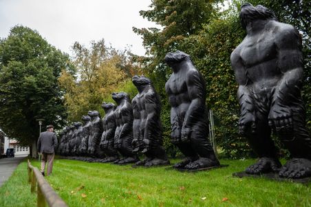 Eine Reihe von großen, schwarzen Affenskulpturen steht im Freien. Ein Mann geht an ihnen vorbei. Die Skulpturen sind in einer Linie aufgestellt.