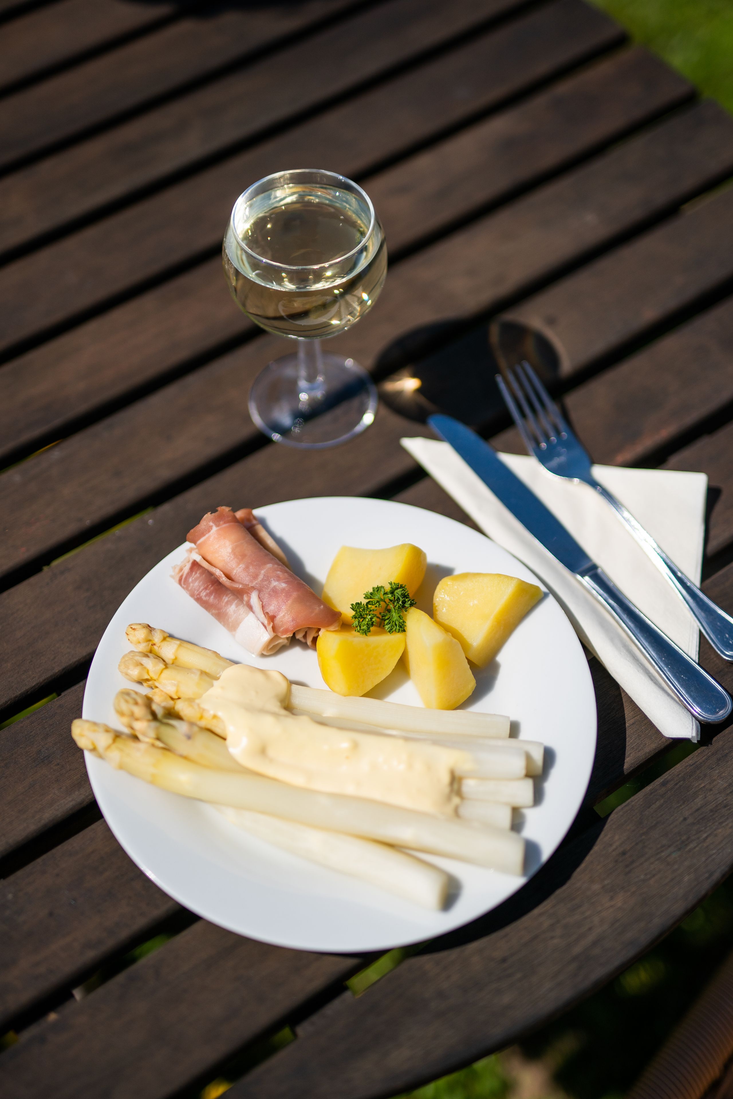 Ein Teller mit Spargel, Schinken, Kartoffeln und Sauce, serviert mit einem Glas Weißwein auf einem Holztisch.