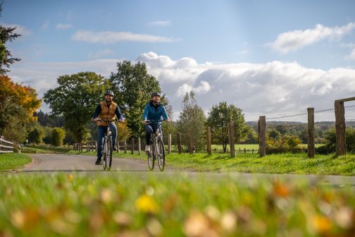 Zwei Radfahrer genießen eine Fahrt auf einem ländlichen Weg, umgeben von grüner Landschaft und herbstlichen Bäumen. Der Himmel ist teils bewölkt.