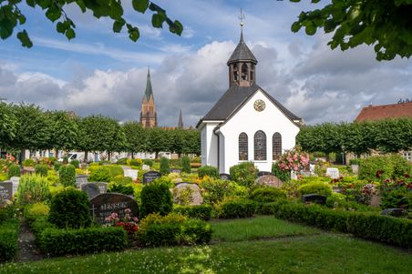 Kleine weiße Friedhofskapelle in Schleswig-Holstein, umgeben von gepflegten Gräbern und blühenden Pflanzen, mit dem St. Petri Dom im Hintergrund.