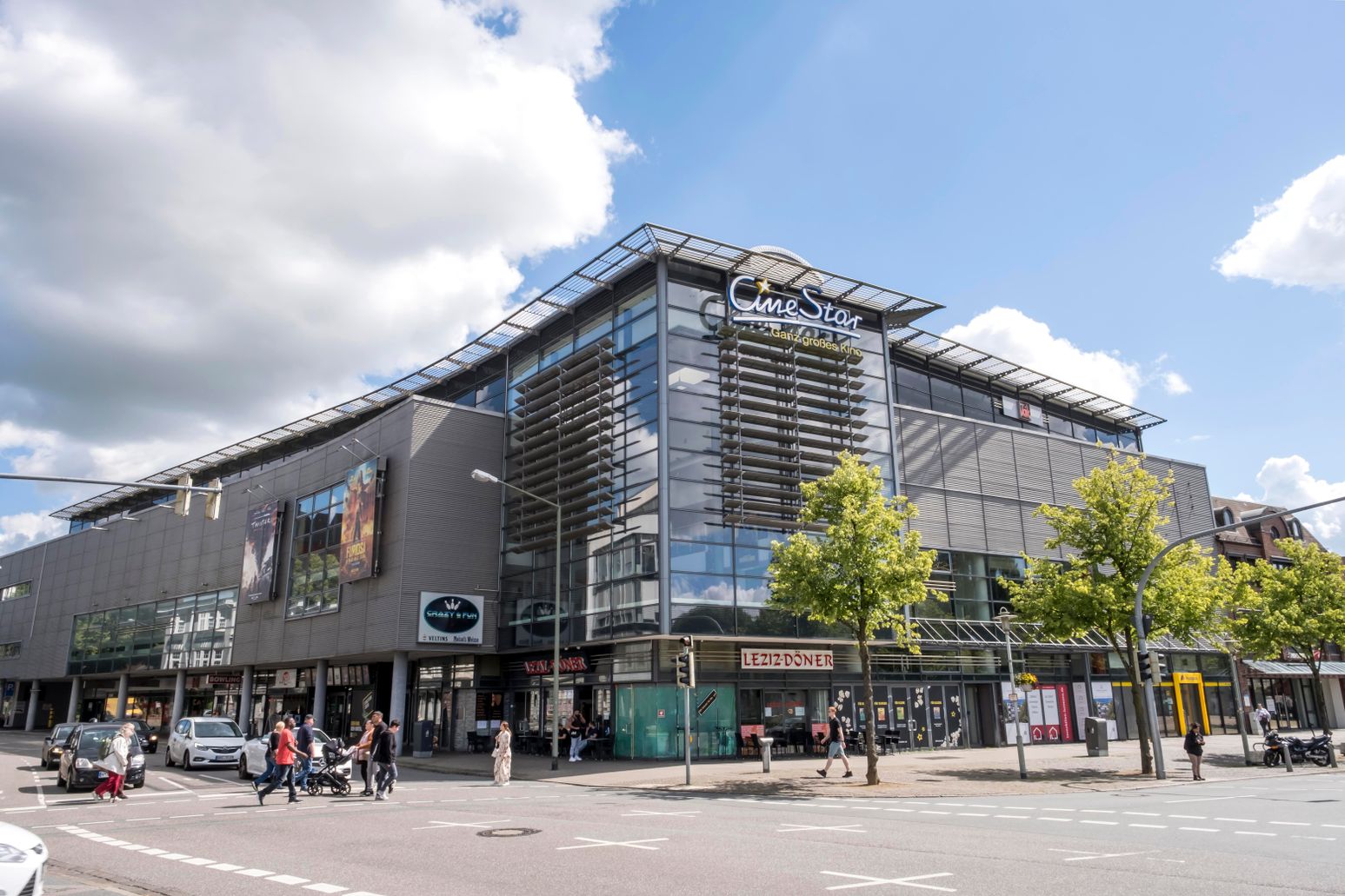 Das CineStar Kino in Neumünster zeigt eine moderne Fassade mit großen Glasflächen. Passanten und Autos beleben die Straßenszene.