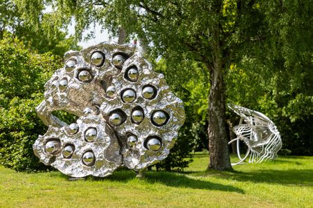 Eine glänzende, metallische Skulptur mit kugelförmigen Elementen steht auf einer Wiese, umgeben von Bäumen. Im Hintergrund ist eine weitere abstrakte Skulptur zu sehen.