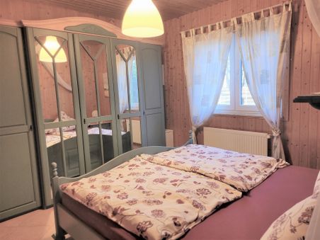 Gemütliches Schlafzimmer mit Holzvertäfelung, Doppelbett, Kleiderschrank mit Spiegeln und Fenster mit hellen Vorhängen.