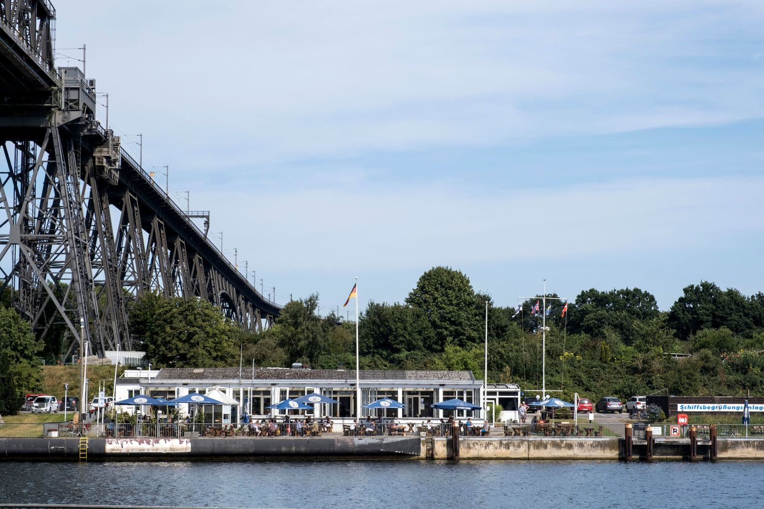 Das Restaurant Brückenterassen liegt malerisch am Wasser, direkt neben einer imposanten Eisenbahnhochbrücke. Gäste sitzen unter Sonnenschirmen.