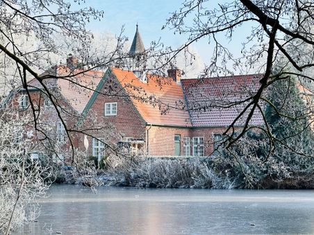 Ein Backsteingebäude mit roten Ziegeldächern, umgeben von frostbedeckten Bäumen und einem zugefrorenen Teich im Vordergrund. Winterliche Atmosphäre.