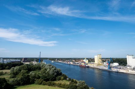 Der Nord-Ostsee-Kanal erstreckt sich unter einem klaren blauen Himmel, gesäumt von Industrieanlagen und grüner Vegetation.