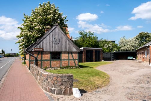 Das Schmiedemuseum Jevenstedt zeigt ein traditionelles Fachwerkgebäude mit Hufeisen-Emblem. Ein gepflegter Garten und eine gepflasterte Einfahrt sind sichtbar.