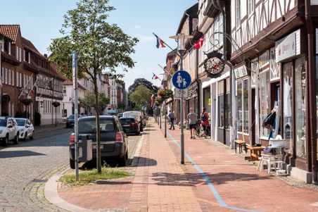 Die Königsstraße in Rendsburg zeigt eine blaue Linie auf dem Gehweg, gesäumt von Fachwerkhäusern und geparkten Autos. Menschen spazieren entlang der Geschäfte.